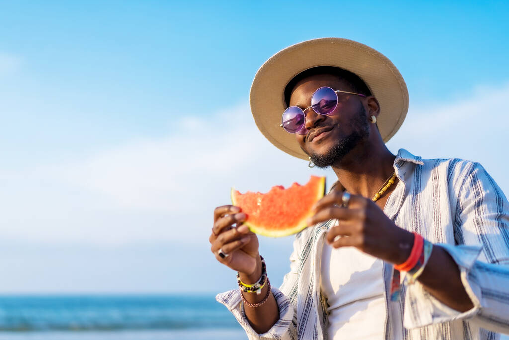 czarny etniczny człowiek cieszyć letnie wakacje na plaży jedząc arbuza z parasolem i okularami - Zdjęcie, obraz