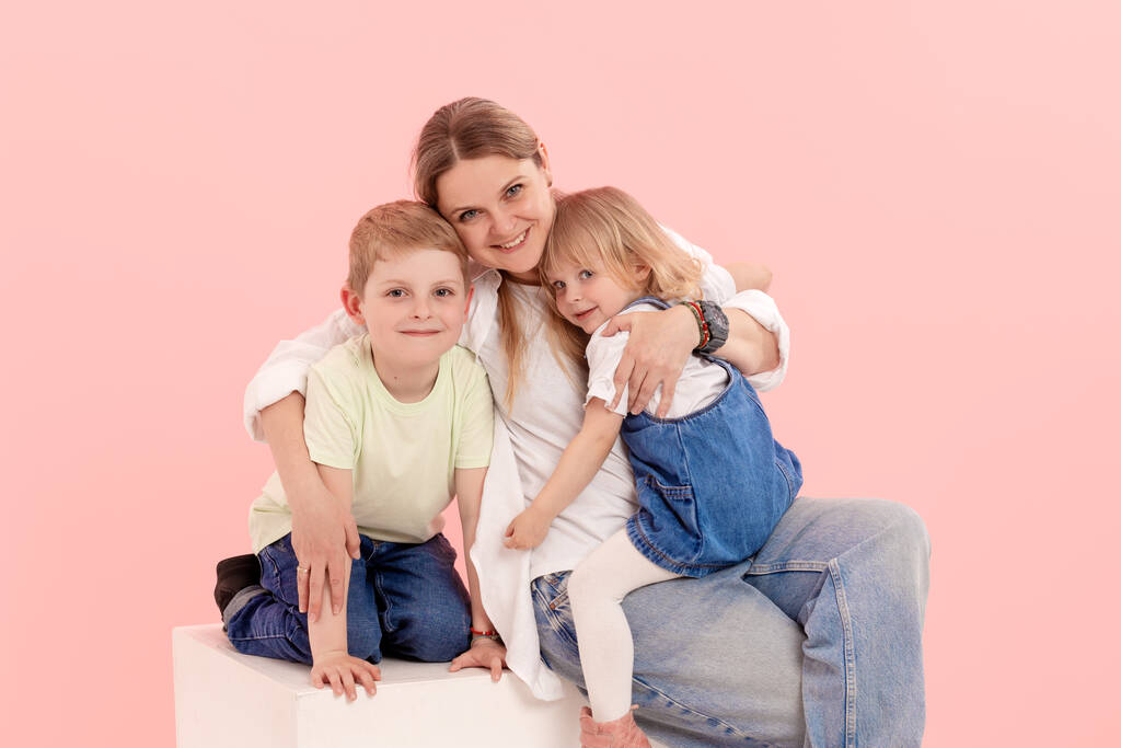 Una joven madre se divierte jugando con una linda hija y un hijo, sosteniendo en sus brazos, abrazándose suavemente sobre un fondo rosa - Foto, imagen