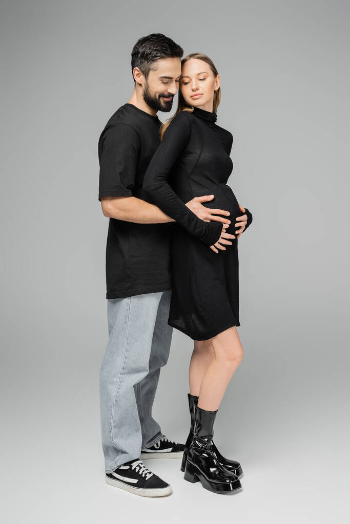 Full length of smiling bearded man in jeans and t-shirt hugging stylish and pregnant wife in black dress while standing together on grey background, growing new life concept - Photo, Image