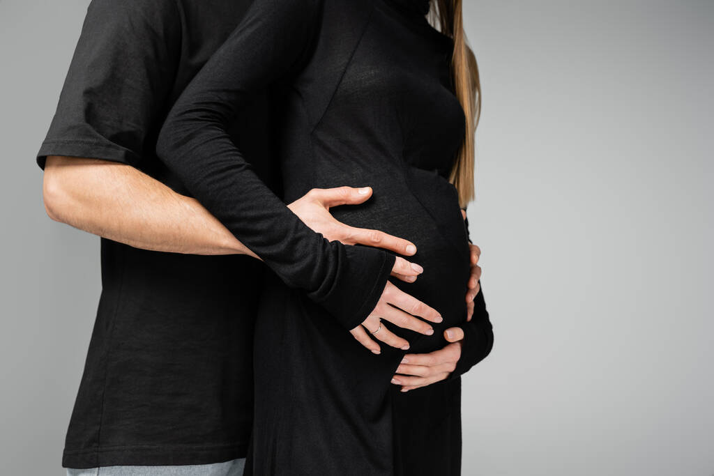 Περικοπή άποψη της φροντίδας άνθρωπος σε μαύρο t-shirt αγκαλιάζει την κοιλιά του κομψό έγκυος γυναίκα στο φόρεμα και στέκεται μαζί απομονώνονται σε γκρι, αυξανόμενη νέα έννοια της ζωής - Φωτογραφία, εικόνα