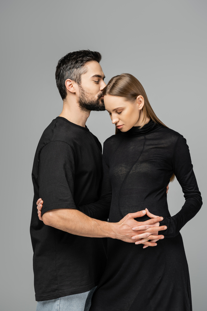Γενειοφόρος σύζυγος σε t-shirt φιλιά και συγκινητική κοιλιά του κομψό έγκυος γυναίκα σε μαύρο φόρεμα και στέκεται απομονωμένη σε γκρι, έννοια της αναμονής των γονέων, ευτυχισμένη οικογένεια  - Φωτογραφία, εικόνα