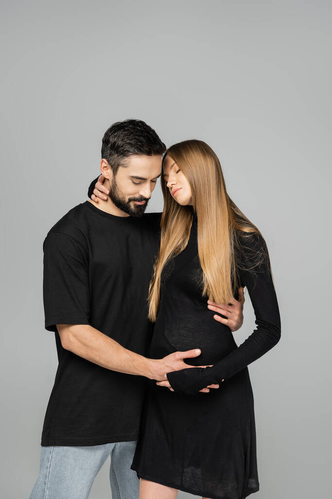 Bärtiger Mann in T-Shirt und Jeans, der den Bauch einer eleganten und entspannten schwangeren Frau berührt, in einem Kleid isoliert auf grau, Neuanfänge und werdende Eltern  - Foto, Bild