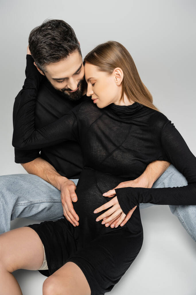 Trendige und schwangere blondhaarige Frau in schwarzem Kleid umarmt bärtigen Ehemann, während sie zusammen auf grauem Hintergrund sitzt, Neuanfänge und Erziehungskonzept  - Foto, Bild