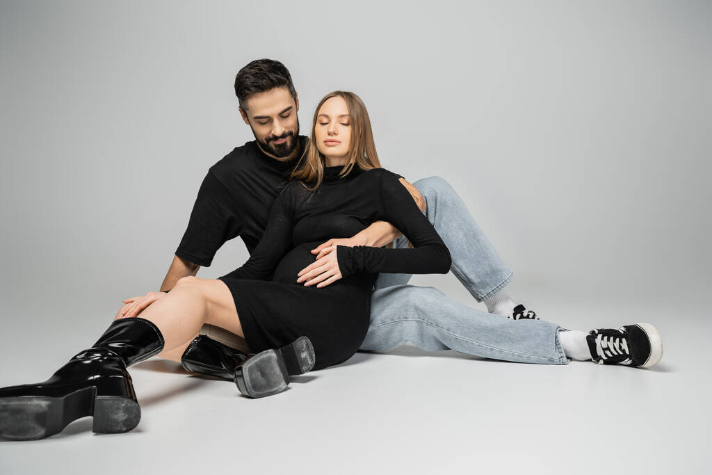 Bearded man in jeans and t-shirt touching belly of fair haired and stylish pregnant wife in black dress while sitting on grey background, new beginnings and parenting concept  - Photo, Image