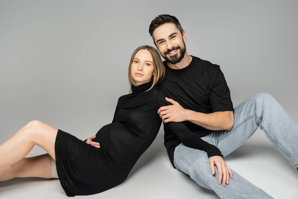 Cheerful husband in t-shirt and jeans looking at camera and hugging trendy pregnant woman in dress while sitting on grey background, new beginnings and parenting concept, husband and wife  - Photo, Image