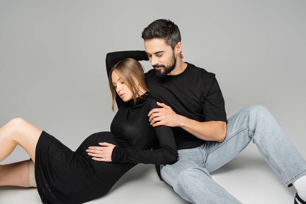 ジーンズに身を包んだ男と黒のTシャツ黒のドレスに身を包んだスタイリッシュで妊娠中の女性がグレーの背景に座って、新しい始まりと子育てのコンセプト、夫と妻  - 写真・画像