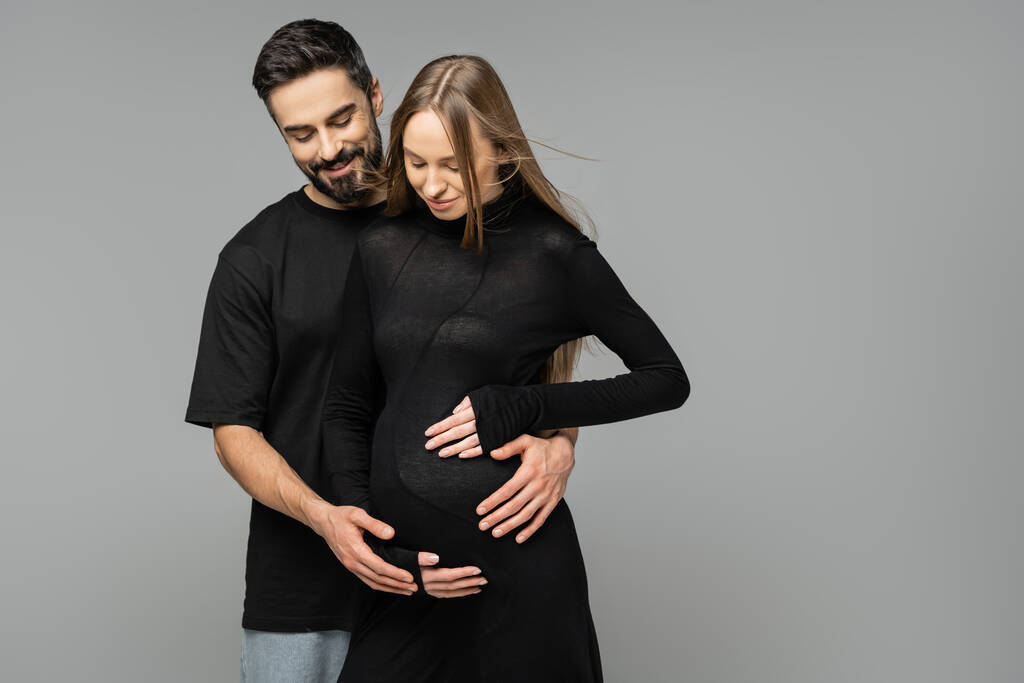 Pozitivní vousatý muž objímání a při pohledu na břicho těhotné ženy ve stylových a černých šatech, zatímco stojí izolované na šedé, nové začátky a očekávání koncepce   - Fotografie, Obrázek
