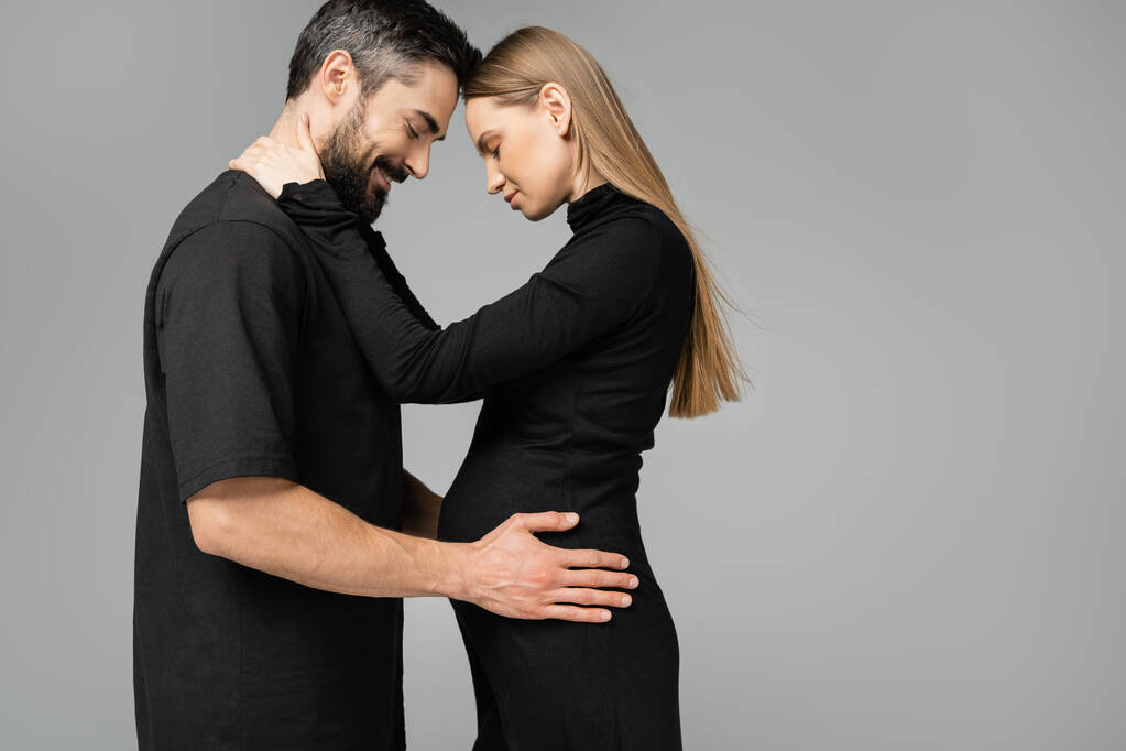 Side view of stylish and fair haired pregnant woman in black dress hugging cheerful husband with closed eyes and standing isolated on grey, new beginnings and anticipation concept   - Photo, Image