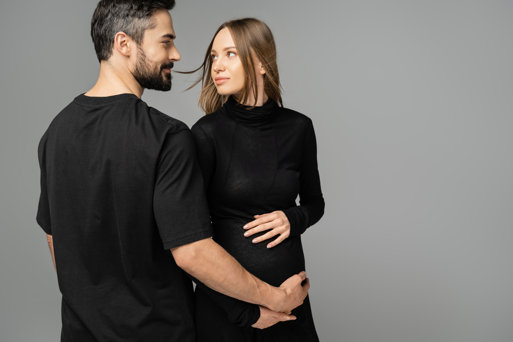 Κομψό και έγκυος γυναίκα με μαύρο φόρεμα αγγίζει την κοιλιά και κοιτάζοντας γενειοφόρος σύζυγος σε t-shirt, ενώ στέκεται απομονωμένη σε γκρι, νέα ξεκινήματα και την έννοια της προσμονής   - Φωτογραφία, εικόνα
