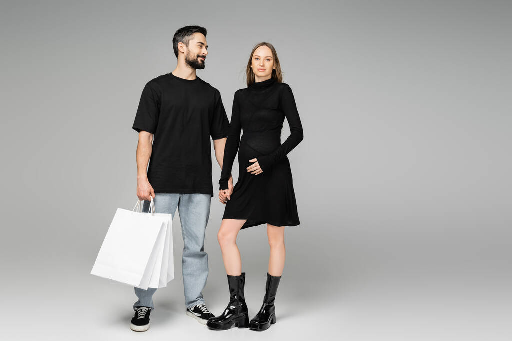 Positive and bearded man holding shopping bags and hand of trendy pregnant wife touching belly and standing together on grey background, new beginnings and shopping concept   - Photo, Image