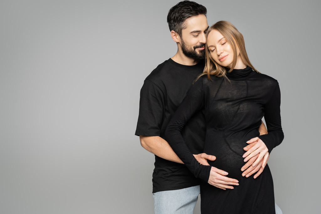 Θετικός γενειοφόρος σύζυγος με μαύρο μπλουζάκι που αγκαλιάζει την κομψή έγκυο γυναίκα με φόρεμα και κλειστά μάτια και στέκεται απομονωμένος σε γκρι, νέα ξεκινήματα και προσμονή   - Φωτογραφία, εικόνα