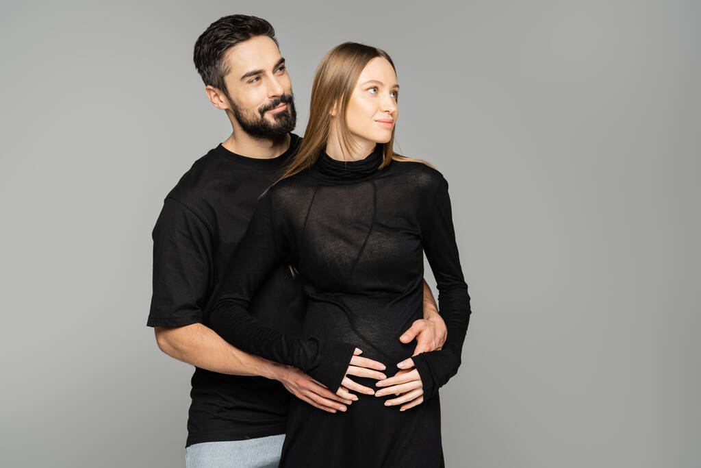 Hymyilevä ja parrakas mies mustassa t-paidassa halaamassa tyylikästä raskaana olevaa vaimoa mekossa ja etsimässä yhdessä eristettynä harmaaseen, uuteen alkuun ja ennakointikonseptiin.   - Valokuva, kuva