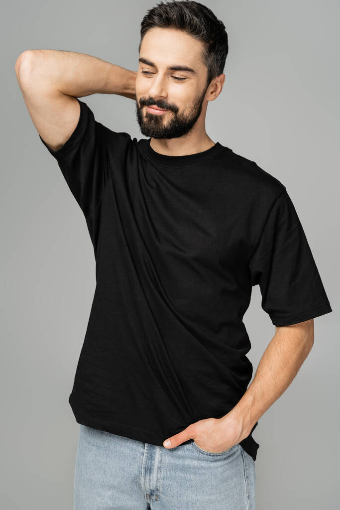 Portrait of relaxed and bearded man in casual black t-shirt and jeans holding hand in pocket and looking away while standing isolated on grey, masculine beauty concept  - Photo, Image