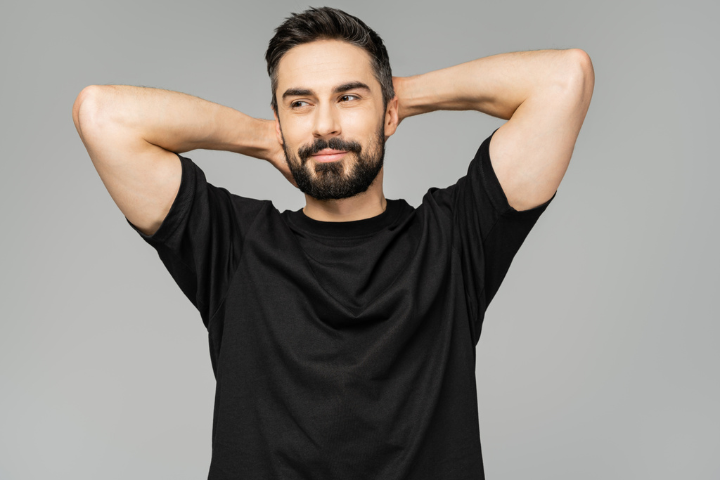 Joyful bearded and brunette man in black t-shirt touching head and looking away while standing isolated on grey, masculine beauty concept, confident and charismatic  - Photo, Image