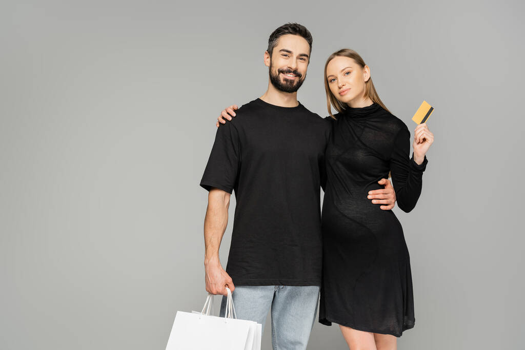 Pozitív szakállas férfi kezében bevásárlótáskák és ölelkező terhes feleség hitelkártyával, miközben a kamerába néz, és elszigetelt szürke, új kezdetek és szülői koncepció   - Fotó, kép