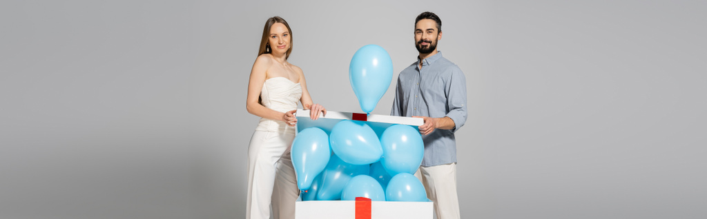 Smiling and trendy expecting parents looking at camera while opening bog gift box with festive blue balloons during gender reveal surprise party isolated on grey, it`s a boy, banner - Photo, Image