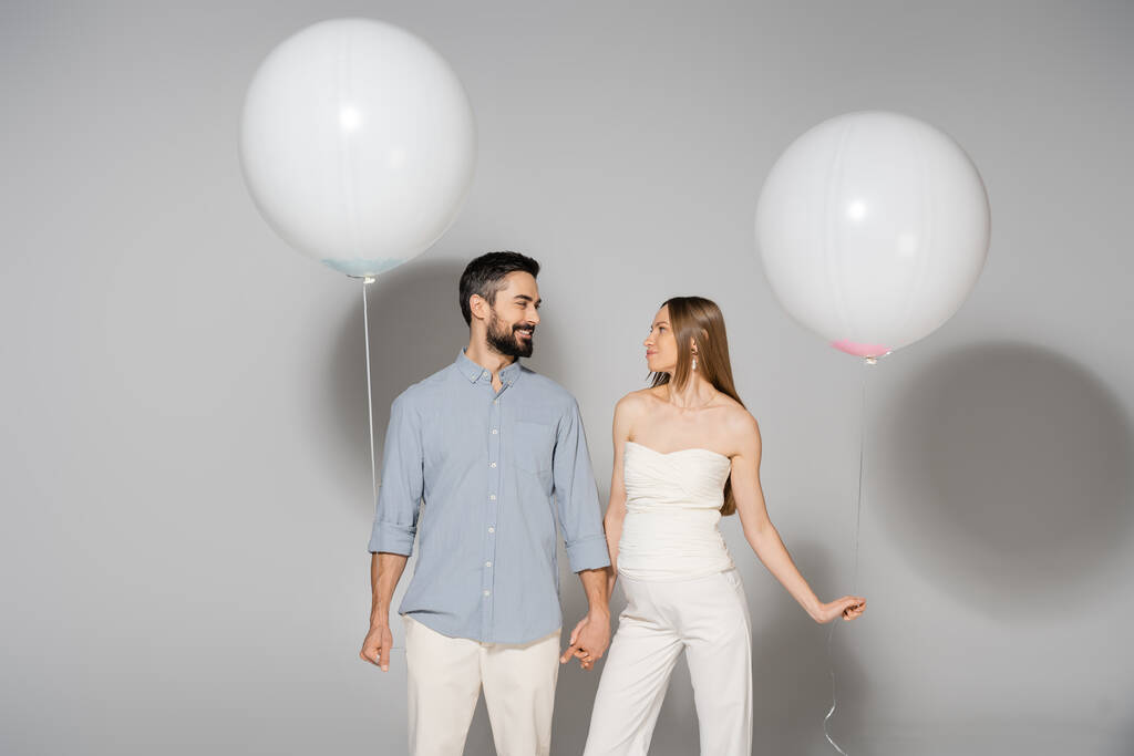 Positive and trendy expecting parents holding hands and festive balloons during celebration and gender reveal surprise party on grey background, expecting parents concept - Photo, Image