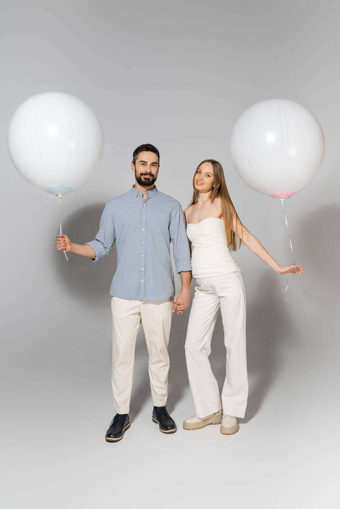 Volledige lengte van modieuze en gelukkig verwachte ouders met witte ballonnen en op zoek naar camera tijdens geslacht onthullen verrassing partij op grijze achtergrond - Foto, afbeelding