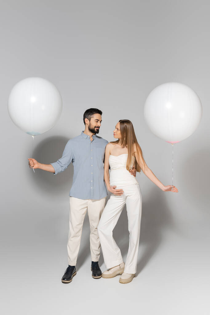 Πλήρες μήκος της μόδας αναμένουν γονείς αγκαλιάζει και κοιτάζοντας ο ένας τον άλλο, ενώ κρατώντας εορταστικά μπαλόνια κατά τη διάρκεια του φύλου αποκαλύπτουν πάρτι έκπληξη σε γκρι φόντο - Φωτογραφία, εικόνα