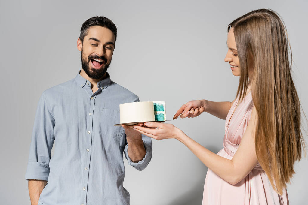 Excited and bearded man holding cake near cheerful pregnant wife with knife during baby shower party on grey background, expecting parents concept, gender party, it`s a boy - Photo, Image