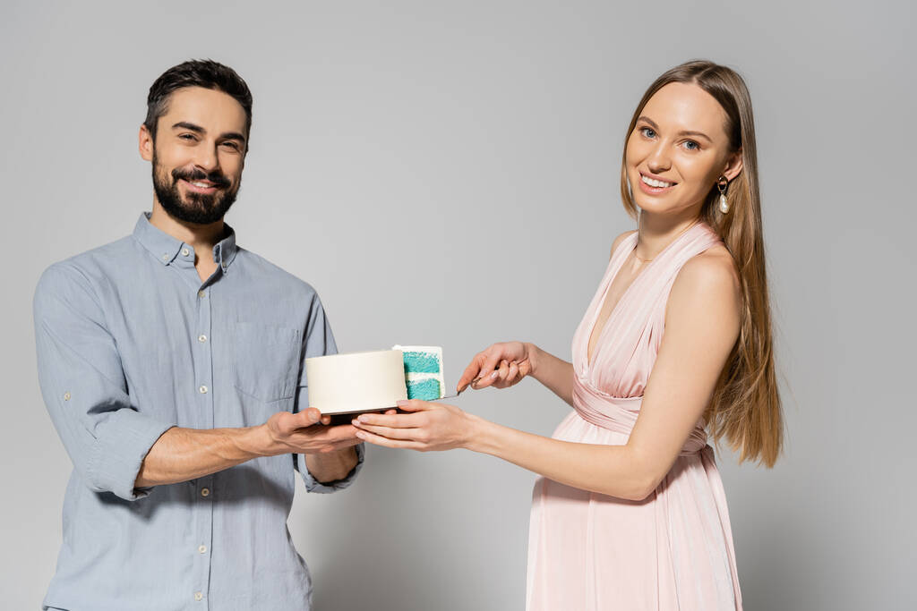 Cheerful and stylish couple looking at camera while cutting blue cake during baby shower celebration on grey background, expecting parents concept, gender party, it`s a boy - Photo, Image