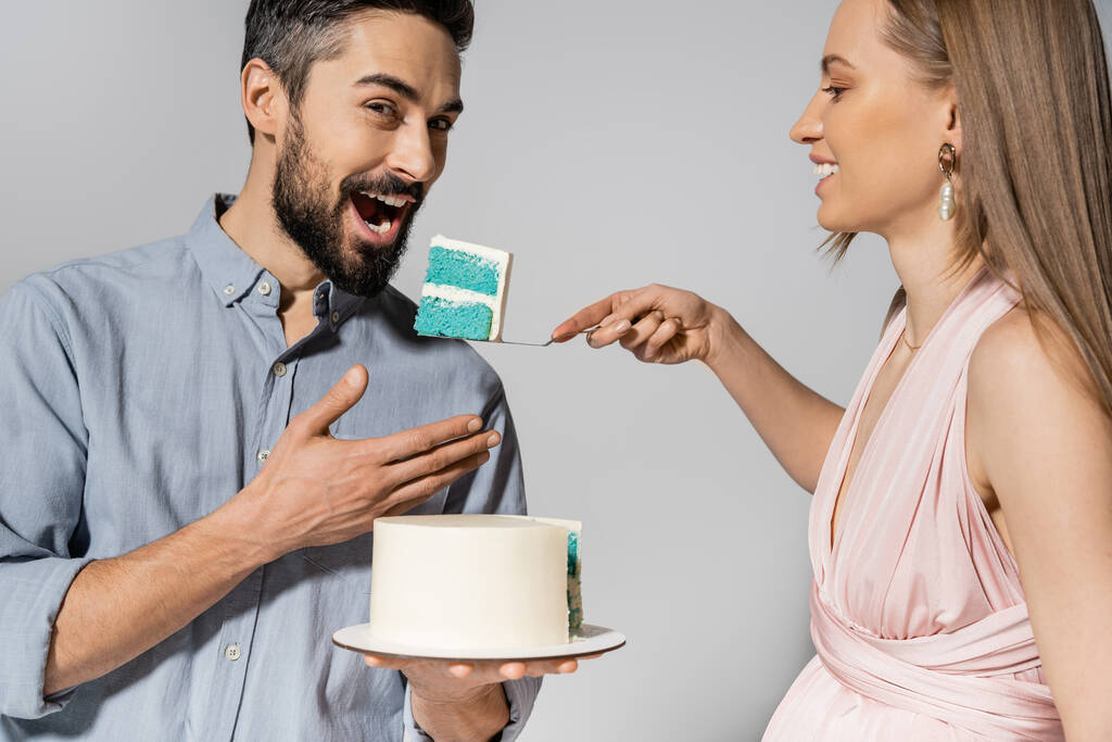 Uśmiechnięta i elegancka kobieta w ciąży karmiąca męża niebieskim tortem podczas obchodów gender party odizolowana na szaro, oczekując rodziców konceptualnego szczęścia, to chłopiec - Zdjęcie, obraz