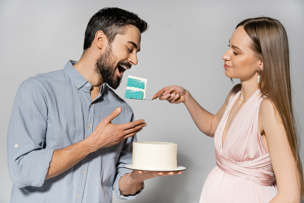 Positieve en elegante zwangere vrouw voeden man met blauwe taart tijdens de baby shower en viering op grijze achtergrond, verwachten ouders concept, gender party, het is een jongen  - Foto, afbeelding