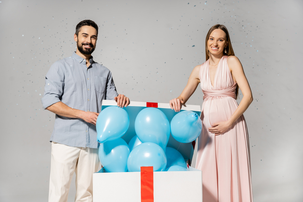Positive pregnant woman in elegant dress opening bog gift box with blue balloons while standing near husband under confetti during baby shower on grey background, gender party, it`s a boy - Photo, Image