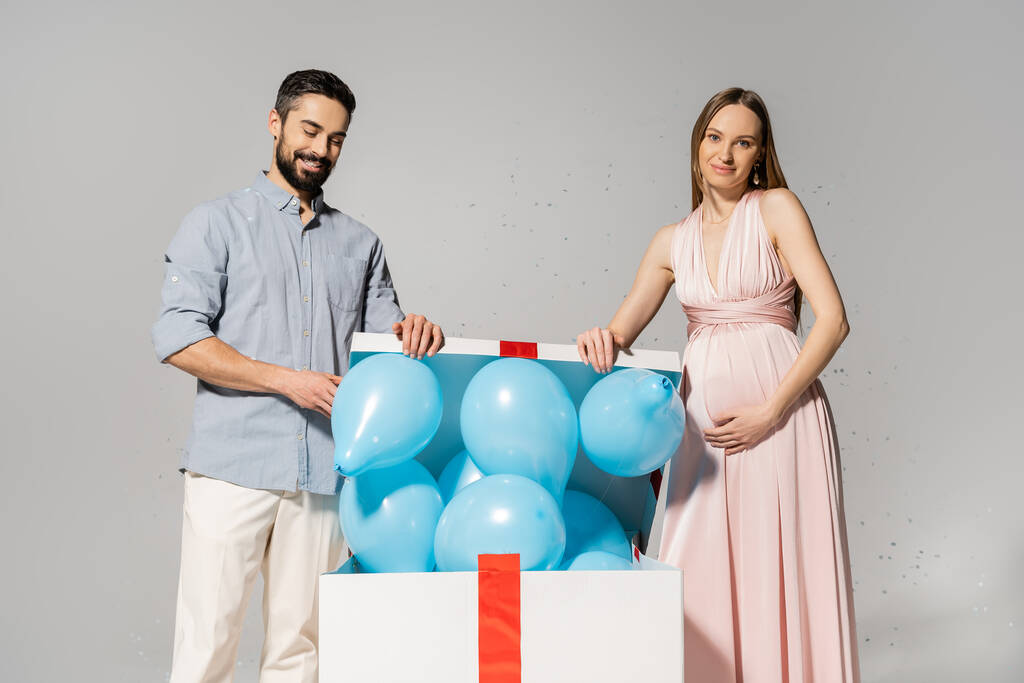 Θετικό και κομψό κουτί δώρου άνοιγμα ζευγάρι με μπλε μπαλόνια κοντά εορταστική κομφετί, ενώ γιορτάζει πάρτι ντους μωρών σε γκρι φόντο, περιμένοντας τους γονείς έννοια, το φύλο κόμμα, είναι ένα αγόρι  - Φωτογραφία, εικόνα