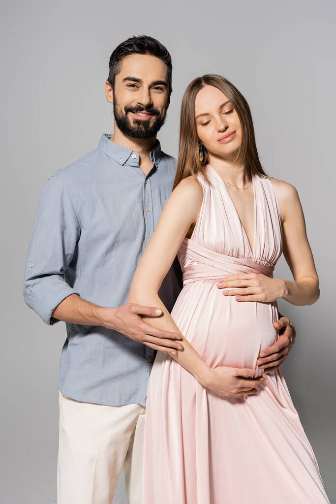 Pozitivní muž objímání elegantní a těhotné ženy v růžových šatech a při pohledu na fotoaparát, zatímco stojí společně na šedém pozadí, očekává rodiče koncept, nové začátky  - Fotografie, Obrázek