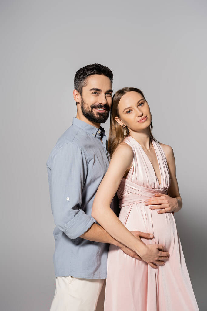 Усміхнений бородатий чоловік обіймає елегантну вагітну жінку в рожевій сукні і дивиться на камеру разом на сірому фоні, очікуючи від батьків концепції, чоловіка і дружини
  - Фото, зображення