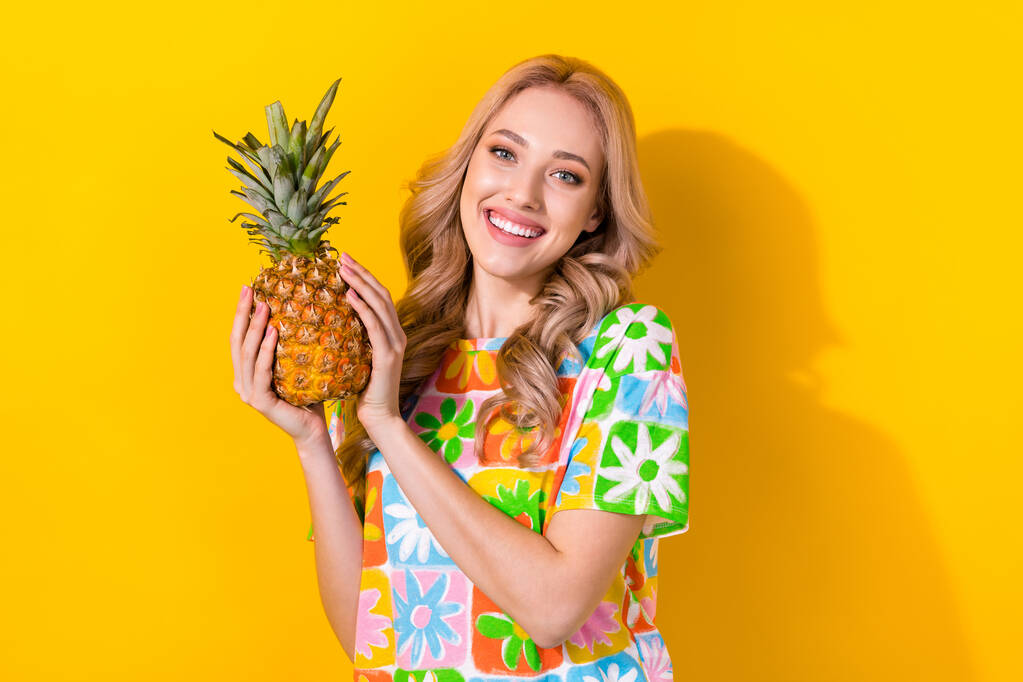 写真楽観的な満足かわいい女の子波状のヘアドレスカラフルなTシャツの手は黄色の色の背景に隔離されたパイナップルを保持. - 写真・画像