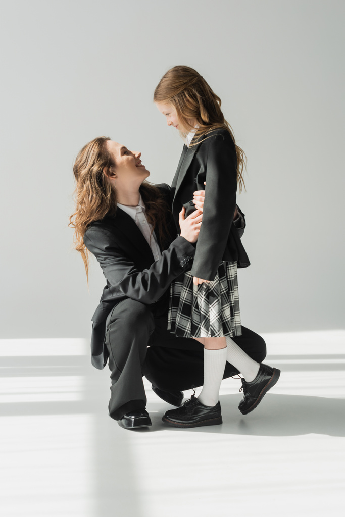 moderne moeder en dochter, zakenvrouw in pak knuffelen schoolmeisje in uniform met geruite rok, blazers, klaar voor nieuw schooljaar, bemoedigend, kijken naar elkaar - Foto, afbeelding