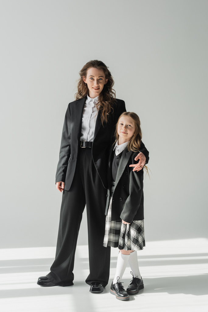 moderne Mutter und Tochter, Geschäftsfrau im Anzug umarmt Schulmädchen in Uniform mit kariertem Rock, auf grauem Hintergrund, Blazer, sich auf das neue Schuljahr vorbereiten, in die Kamera schauen, formelle Kleidung  - Foto, Bild