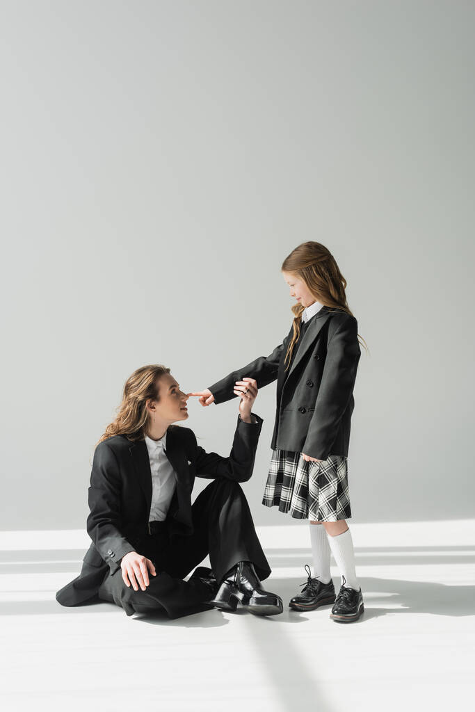 школьница трогает нос работающей матери, веселая девушка в школьной форме смотрит на деловую женщину в костюме на сером фоне, формальный наряд, модную семью, сближение, современное воспитание  - Фото, изображение