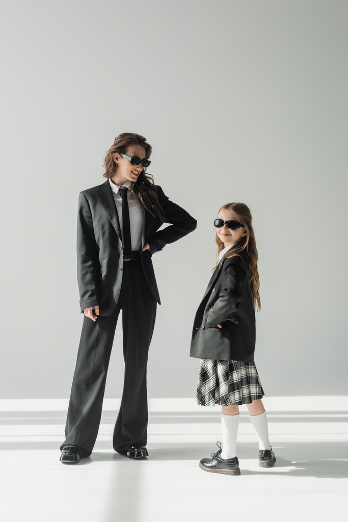 cheerful woman with her daughter, businesswoman in suit posing with hand on hip and schoolgirl in sunglasses and uniform standing together on grey background in studio, formal attire  - Photo, Image