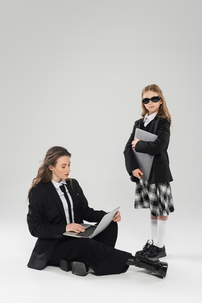 digital nomadism, fashionable woman in suit using laptop near daughter in school uniform and sunglasses on grey background, remote work, working mother, business attire  - Photo, Image