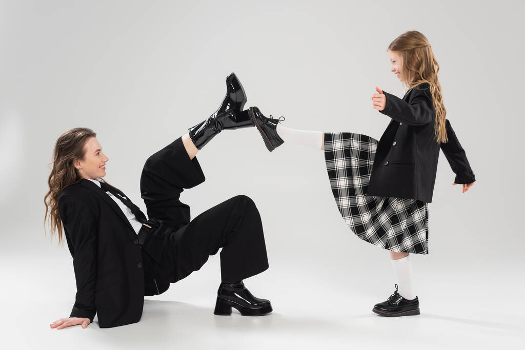 modern parenting, having fun, side view of happy mother in suit and child in school uniform touching boots of each other on grey background in studio, blazer, businesswoman, back to school  - Photo, Image