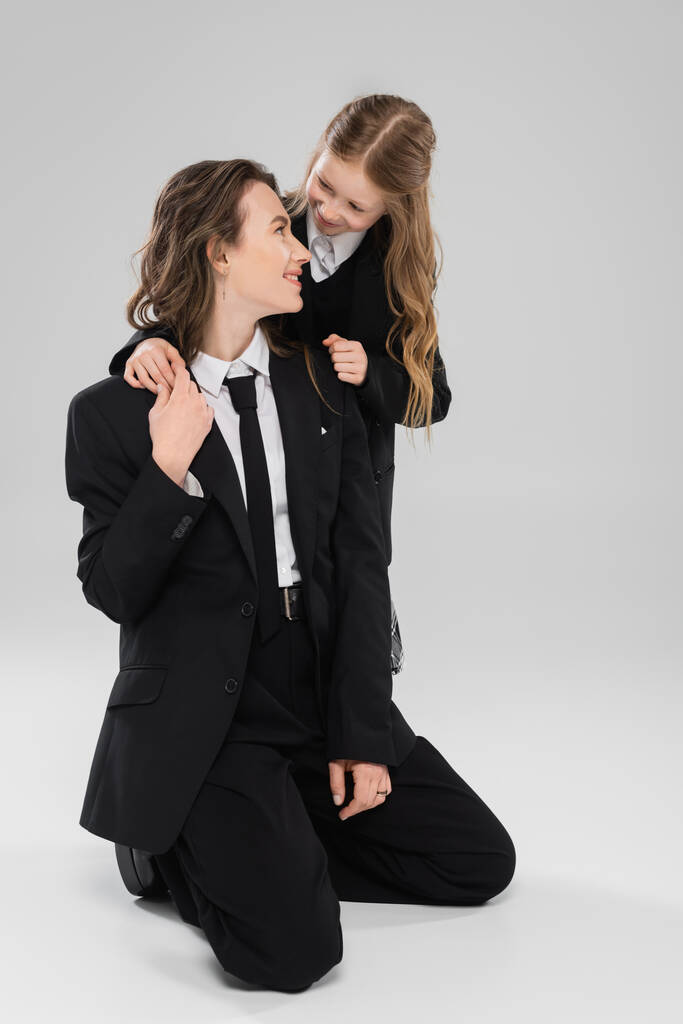 glückliches Schulmädchen umarmt arbeitende Mutter, fröhliches Mädchen in Schuluniform schaut Mama im Anzug auf grauem Hintergrund an, formelle Kleidung, modische Familie, Bindung, moderne Elternschaft  - Foto, Bild