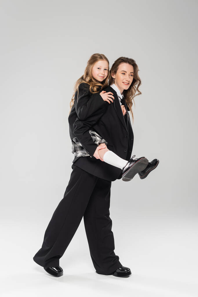 mother piggybacking daughter in school uniform, working mom in business attire and schoolgirl on grey background in studio, modern parenting, fashionable family, having fun  - Photo, Image