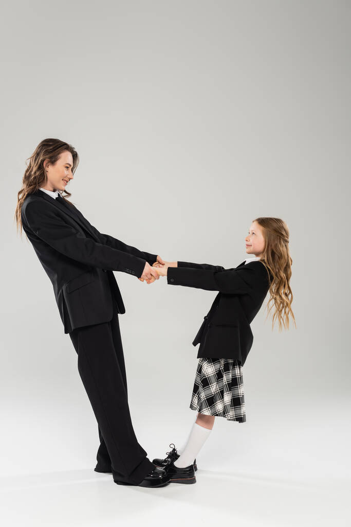 mother and schoolgirl holding hands, happy woman in business attire and child in school uniform standing together on grey background, modern parenting, back to school  - Photo, Image