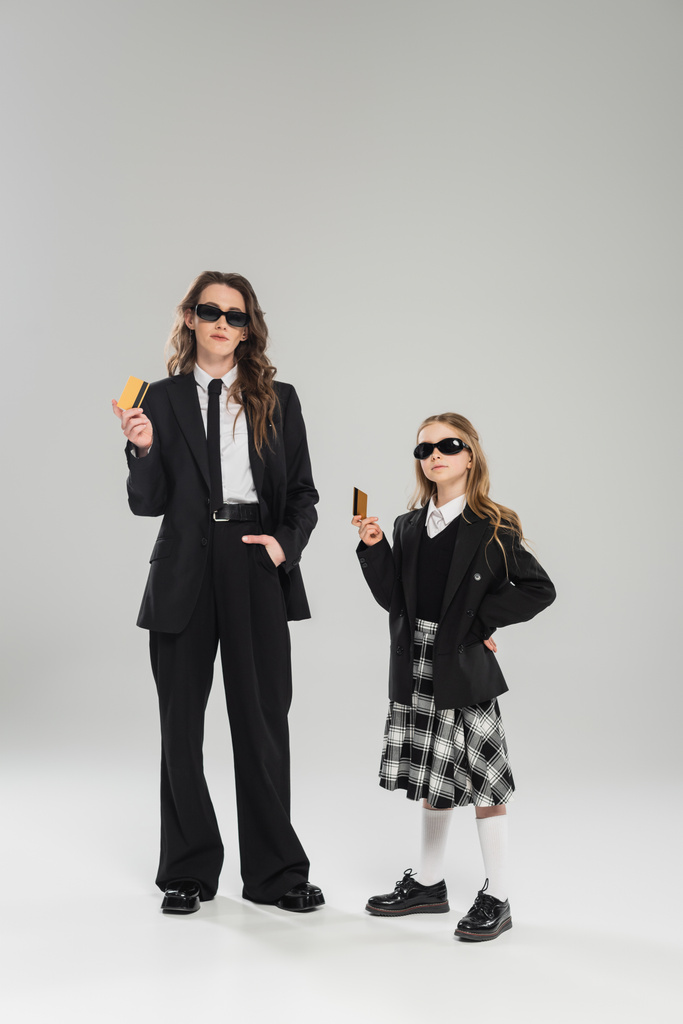 matka a dcera v slunečních brýlích, podnikatelka v obleku a školačka v uniformě držící kreditní karty na šedém pozadí, moderní rodičovství, finanční učení, rozpočtování, správa peněz   - Fotografie, Obrázek