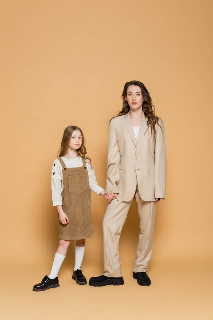 μοντέρνα μητέρα και κόρη, κομψή εργαζόμενη μητέρα με κοστούμι κρατώντας τα χέρια με την κόρη, ενώ στέκεται μαζί σε μπεζ φόντο, γονέας και παιδί, μοντέρνα οικογένεια  - Φωτογραφία, εικόνα