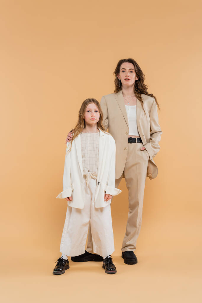 stylish mother and daughter in suits, woman and girl looking at camera while standing together on beige background, fashionable outfits, formal attire, corporate mom, modern family, hand in pocket   - Photo, Image