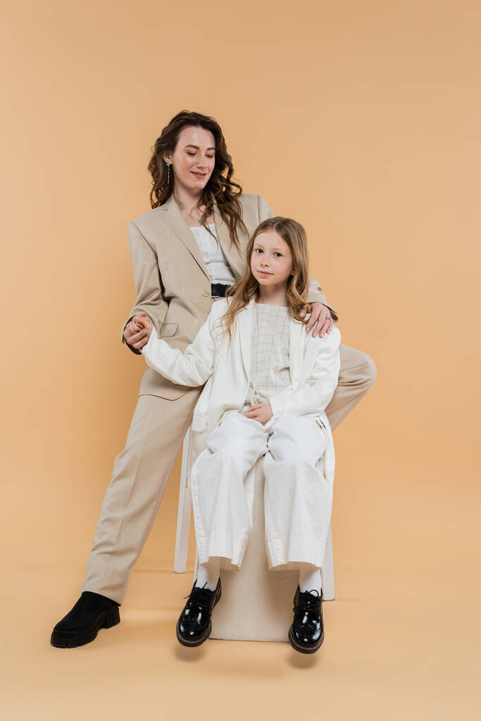 élégant mère et fille en costumes, femme et fille tenant la main et assis ensemble sur fond beige, tenues à la mode, tenue formelle, maman d'entreprise, famille moderne  - Photo, image