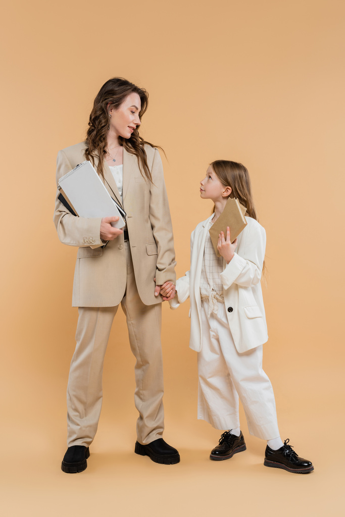 trendy mother and daughter in suits, woman and child holding books and notebooks while standing together on beige background, fashionable outfits, formal attire, corporate mom, education concept  - Photo, Image
