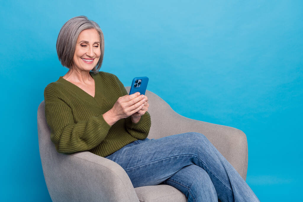 Фото оптимистично удовлетворены приятный человек носить вязаный свитер читать электронную почту на смартфоне сидеть в кресле изолированы на синем фоне цвета. - Фото, изображение