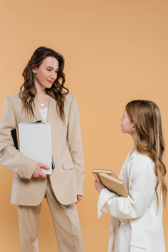 fashionable family, mother and daughter in suits, woman and child holding books and notebooks while standing together on beige background, formal attire, corporate mom, education concept  - Photo, Image