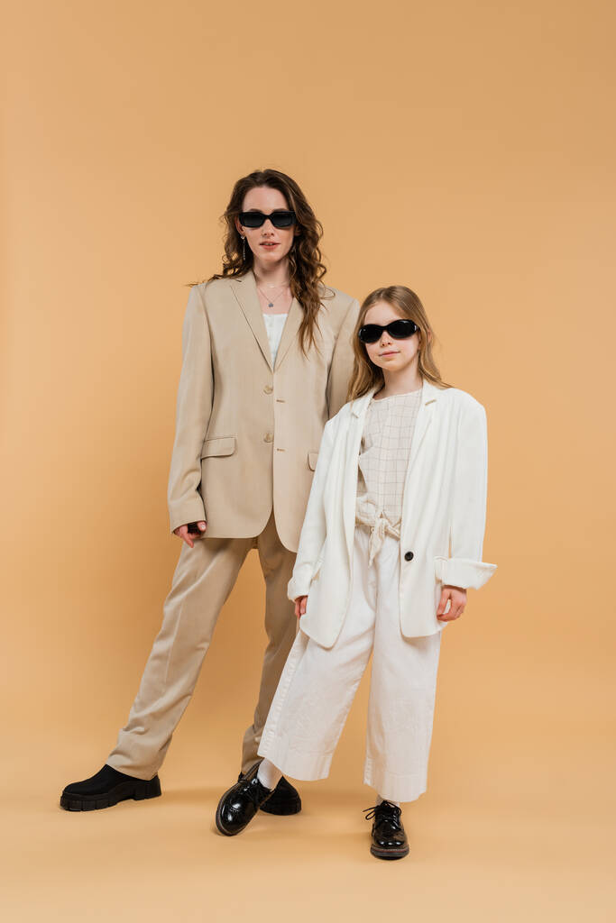 trendy mother and daughter in sunglasses, businesswoman and girl in suits  standing together on beige background, fashionable outfits, formal attire, corporate mom, modern family  - Photo, Image