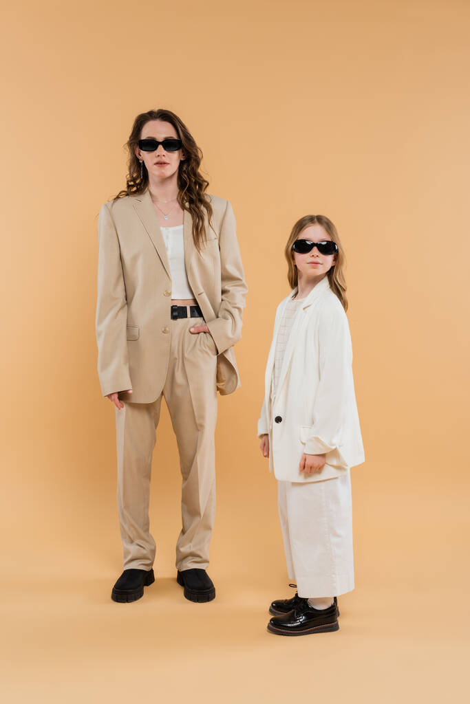 moderne familie, moeder en dochter in zonnebril, stijlvolle zakenvrouw en meisje in kostuums samen poseren op beige achtergrond, modieuze outfits, formele kleding, corporate mam  - Foto, afbeelding